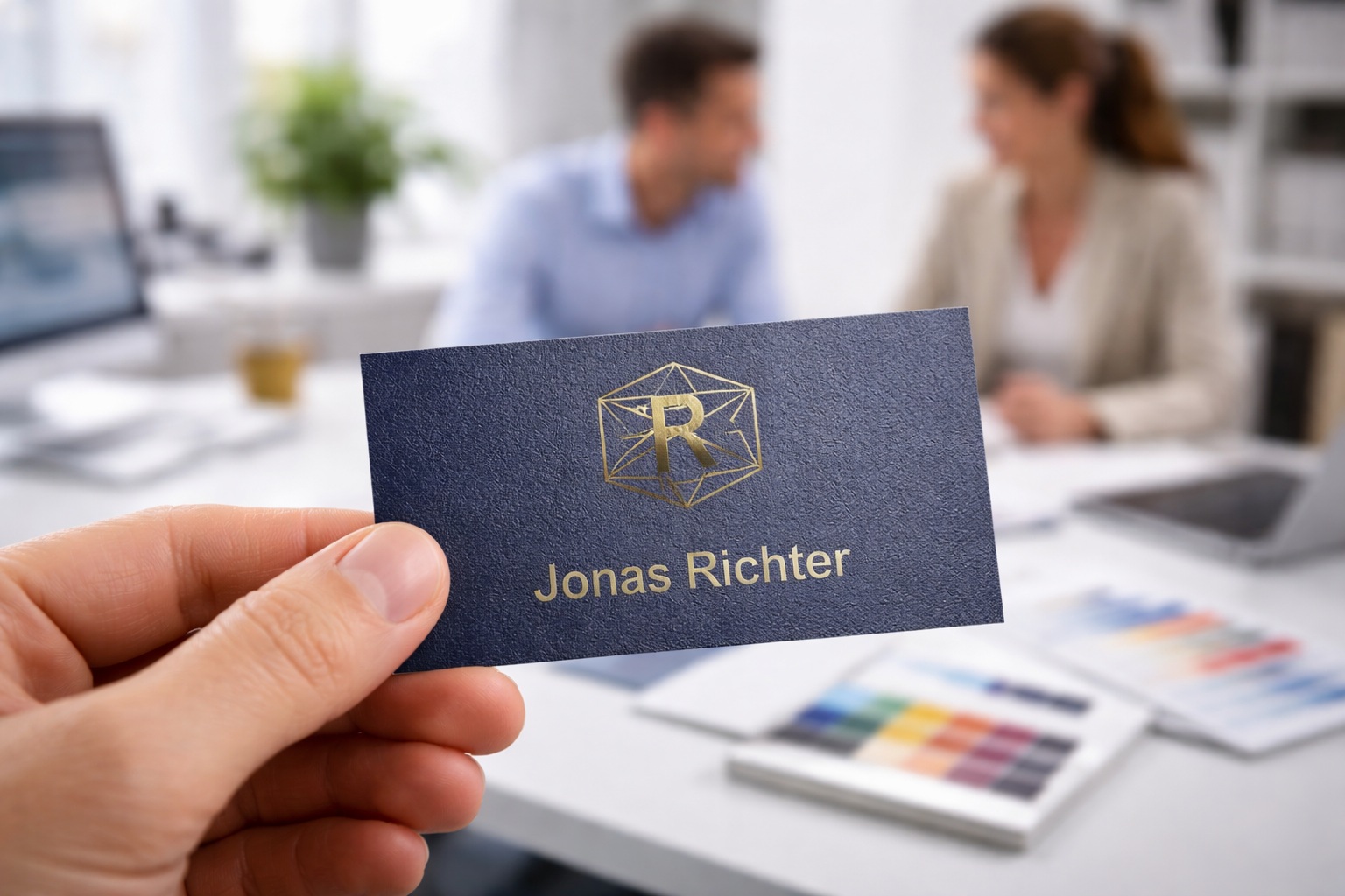 Visitenkarte mit Goldprägung wird in einem Büro gezeigt, im Hintergrund zwei Personen im Gespräch